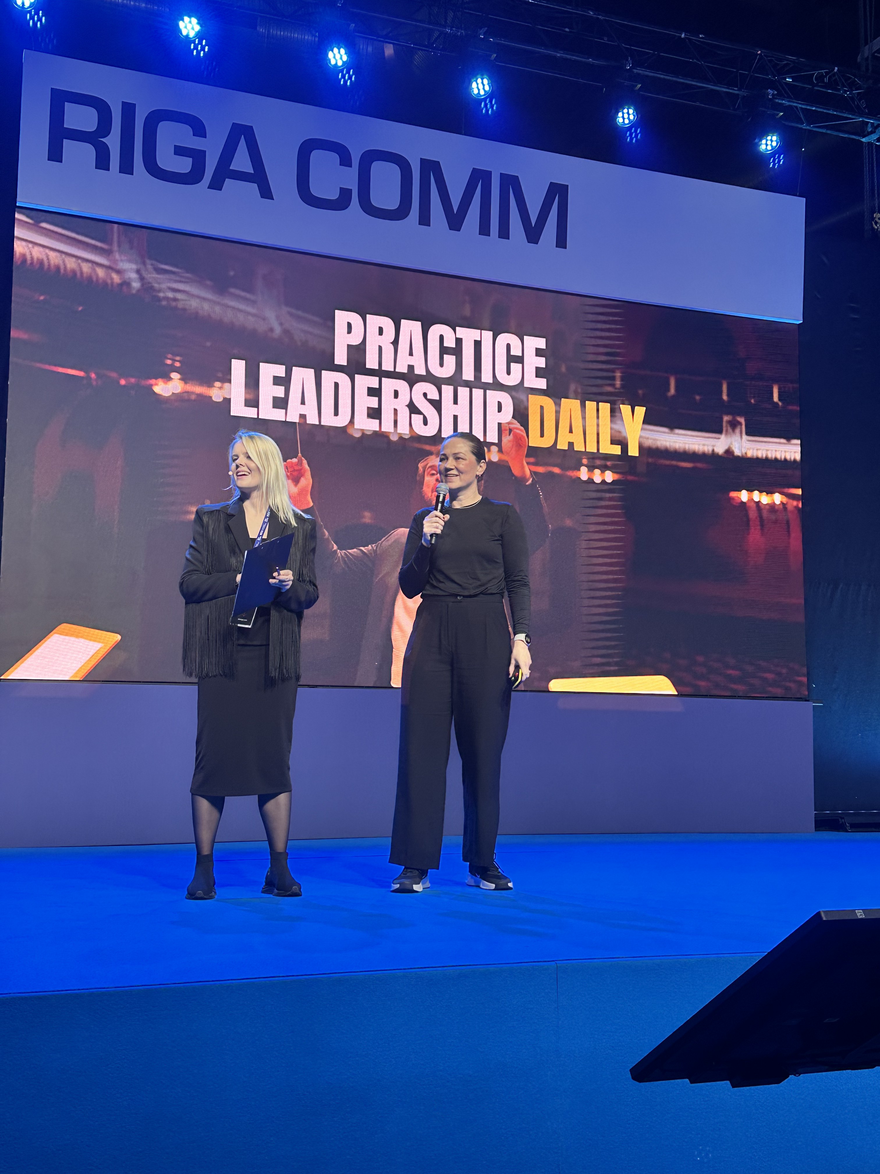 Ieva Drazniece on stage with client Liina Liiv at RIGA COMM, in front of a Practice Leadership Daily slide