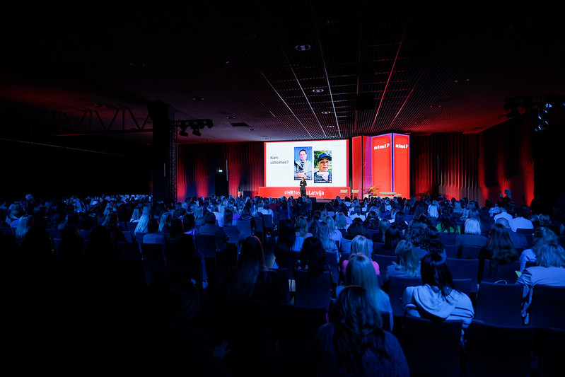 Ieva Drazniece speaking on stage to a packed conference hall, with the question 'Kam uzticēties?' on the screen behind her